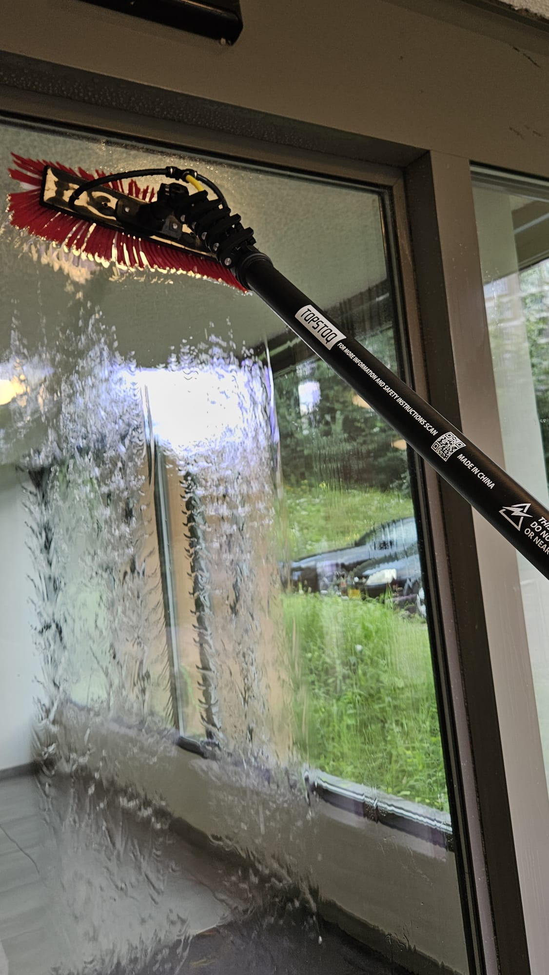 Professionele glazenwasser tuckert een deurraam met de Topstoq carbon telescoopsteel, ingeschoven voor lichtgewicht gebruik en optimale controle bij het borstelen en spoelen.